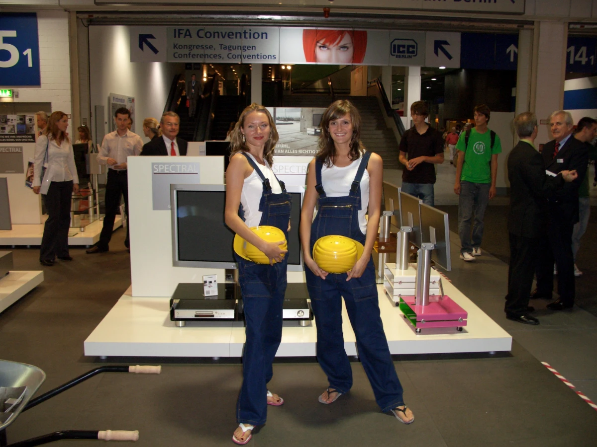 Zwei Frauen mit braunen Haaren in Latzhosen und wei&szlig;en T-Shirts halten gelbe Bauhelme vor einem Messestand, Promotion Team im Einsatz