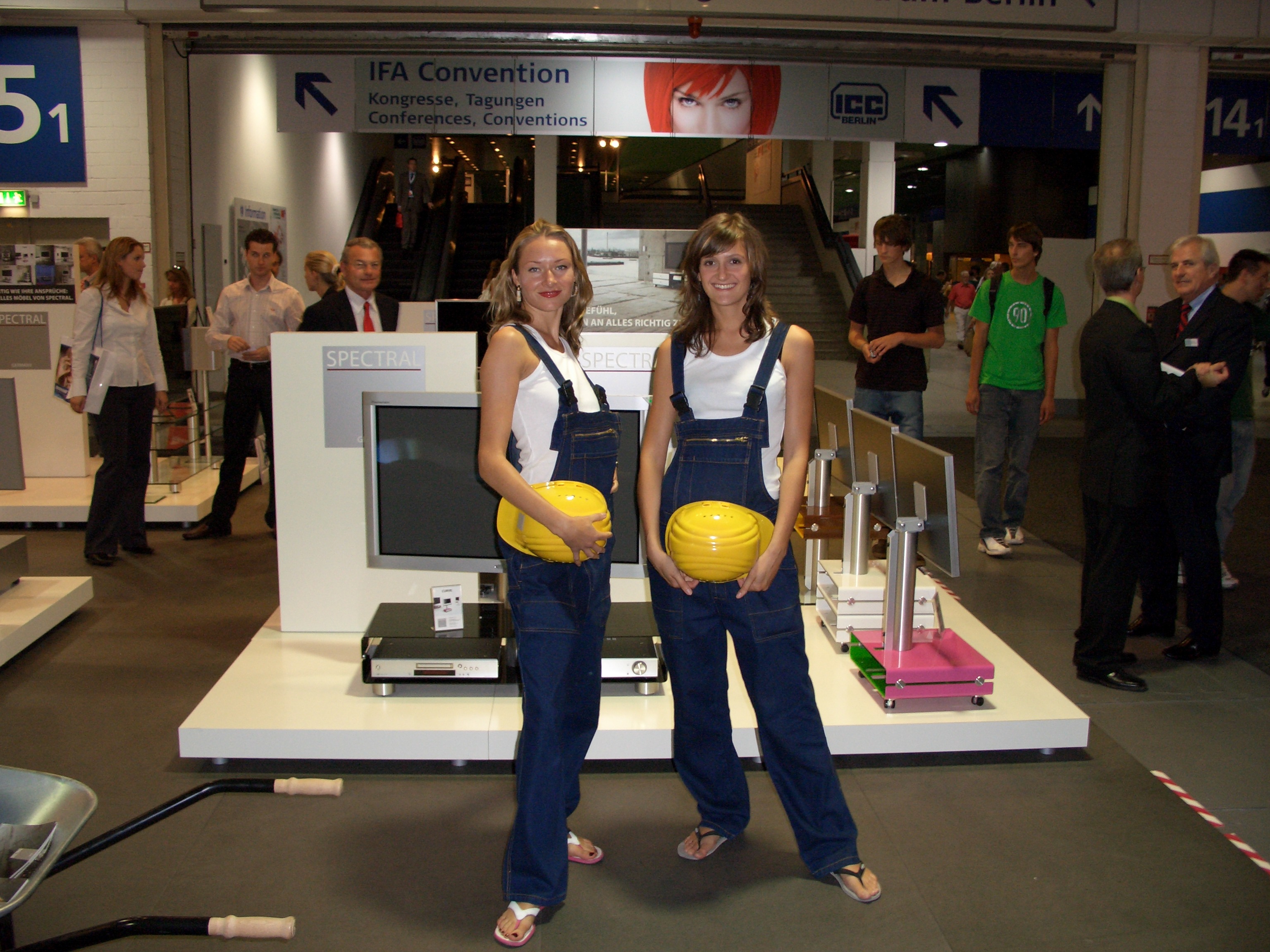 Zwei Frauen mit braunen Haaren in Latzhosen und wei&szlig;en T-Shirts halten gelbe Bauhelme vor einem Messestand, Promotion Team im Einsatz