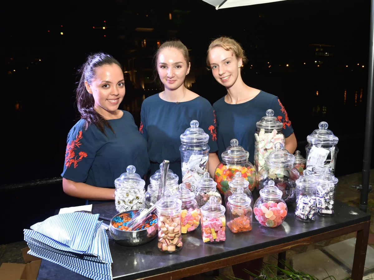 Drei Frauen in dunkelblauen Blusen stehen l&auml;chelnd vor einem S&uuml;&szlig;igkeiten-Tisch bei einem Event, freundliche Hosts im Einsatz bei Promotion und Messe