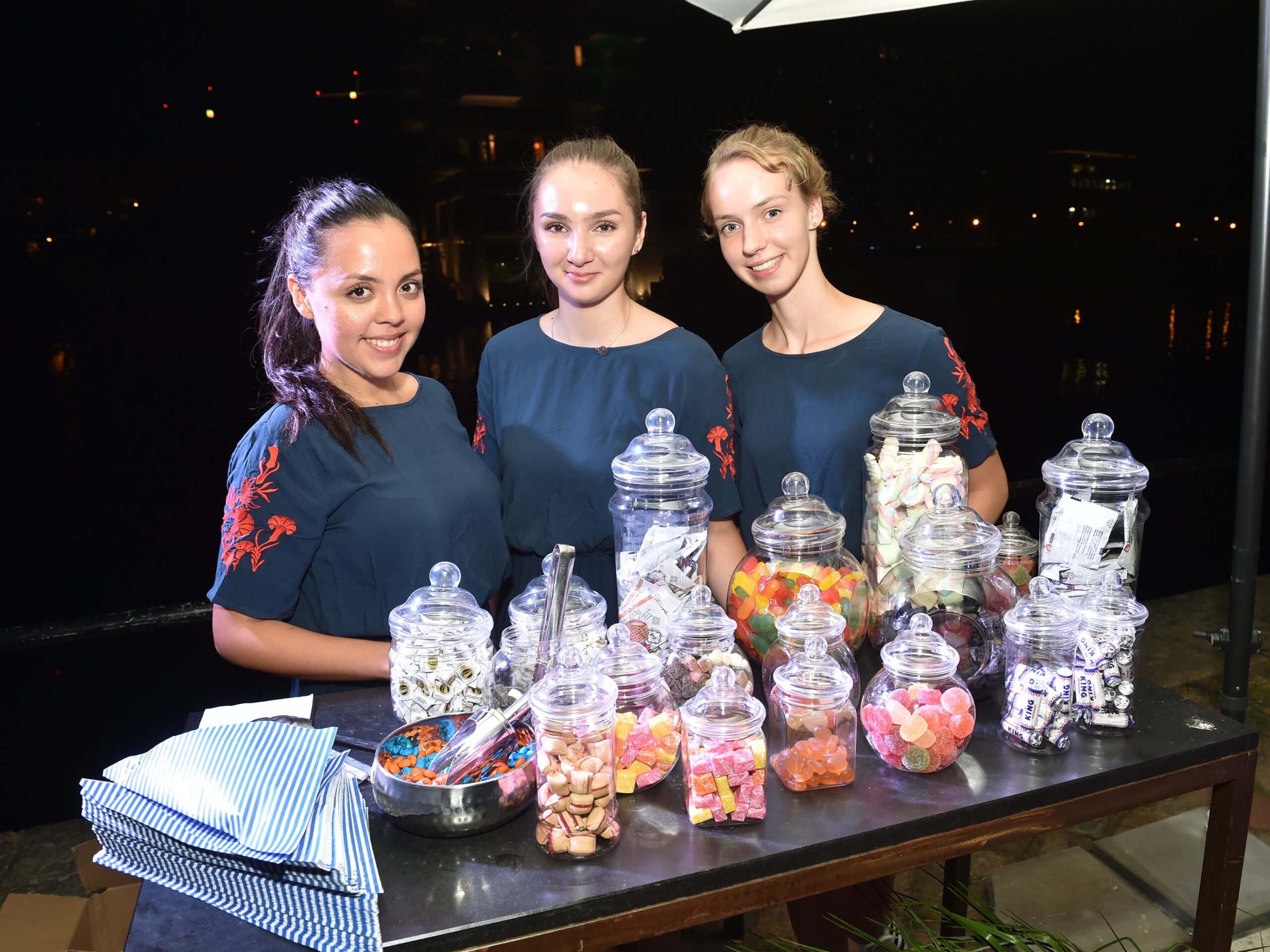 Drei Frauen in dunkelblauen Blusen stehen l&auml;chelnd vor einem S&uuml;&szlig;igkeiten-Tisch bei einem Event, freundliche Hosts im Einsatz bei Promotion und Messe