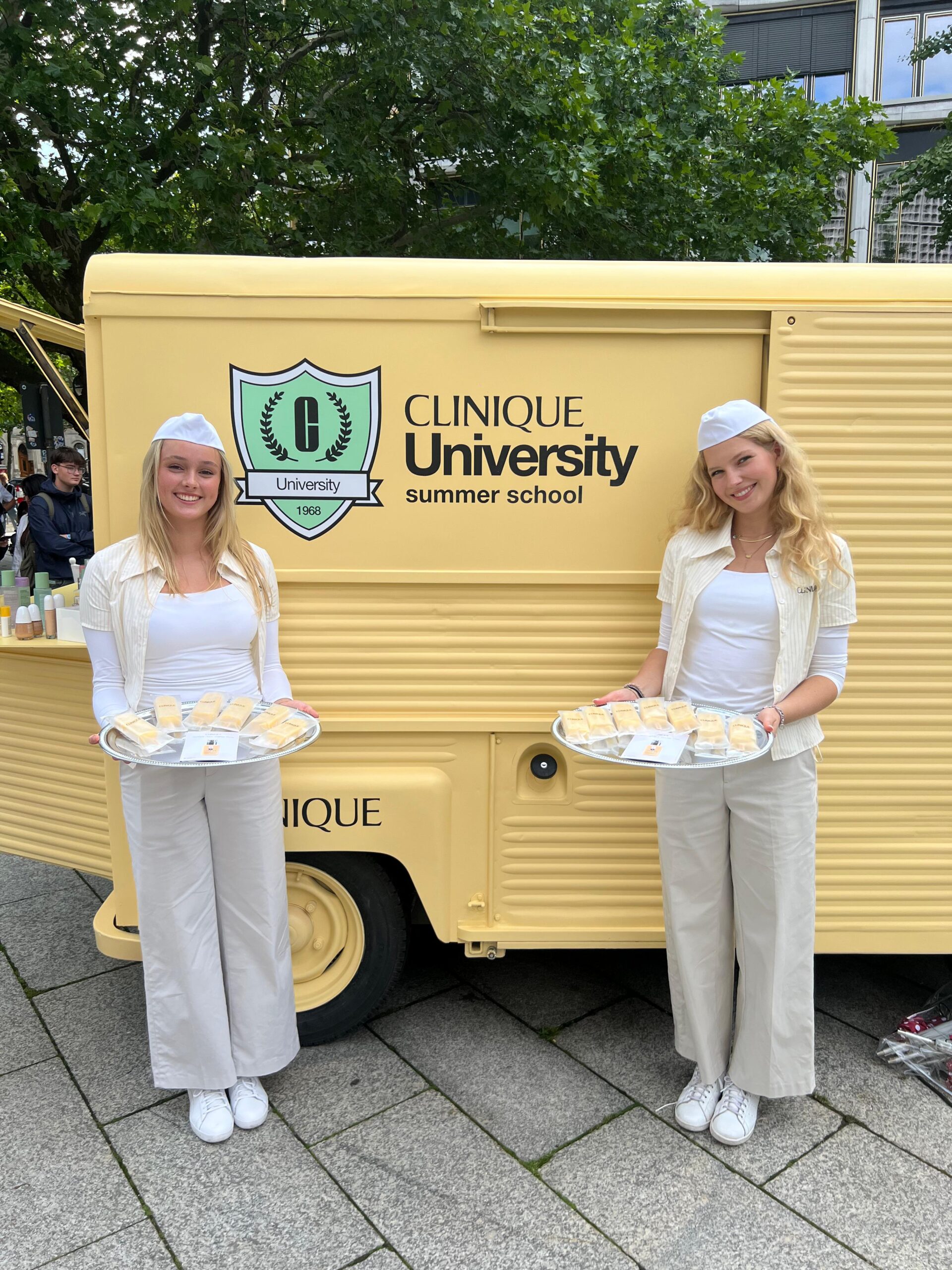 Zwei Promotion Hostessen stehen vor einem gelben Retro-Bus mit Markenbranding, präsentieren Produkte auf Tablets, lächelnder Einsatz bei Event und Markenaktion