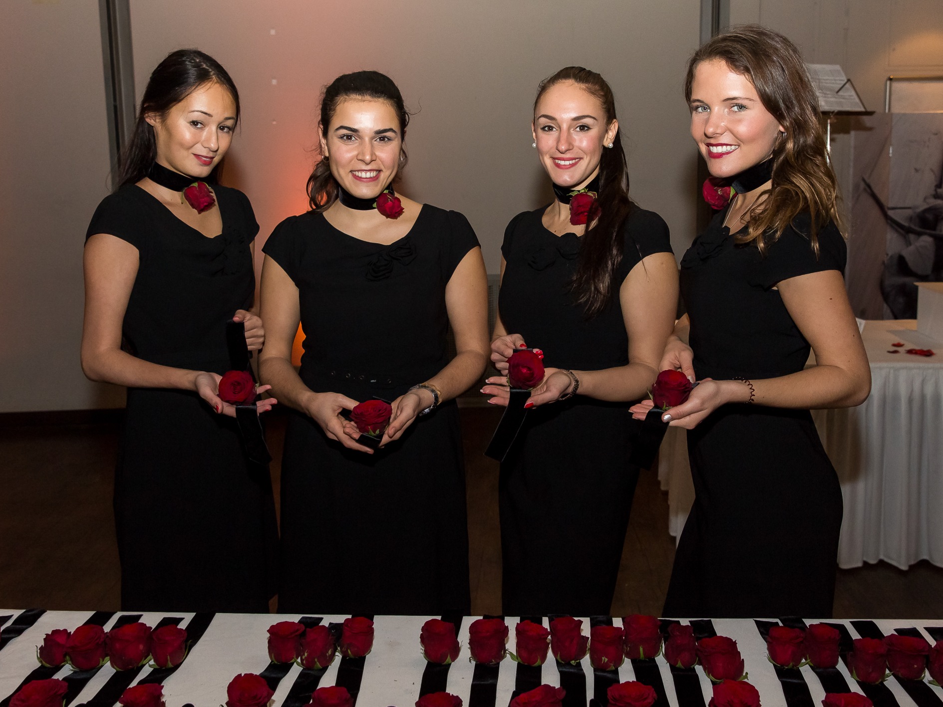 > Vier Hostessen in eleganten schwarzen Kleidern an der Akkreditierung, rote Armbänder auf einem Tisch, professioneller Check-in bei Event und Veranstaltung