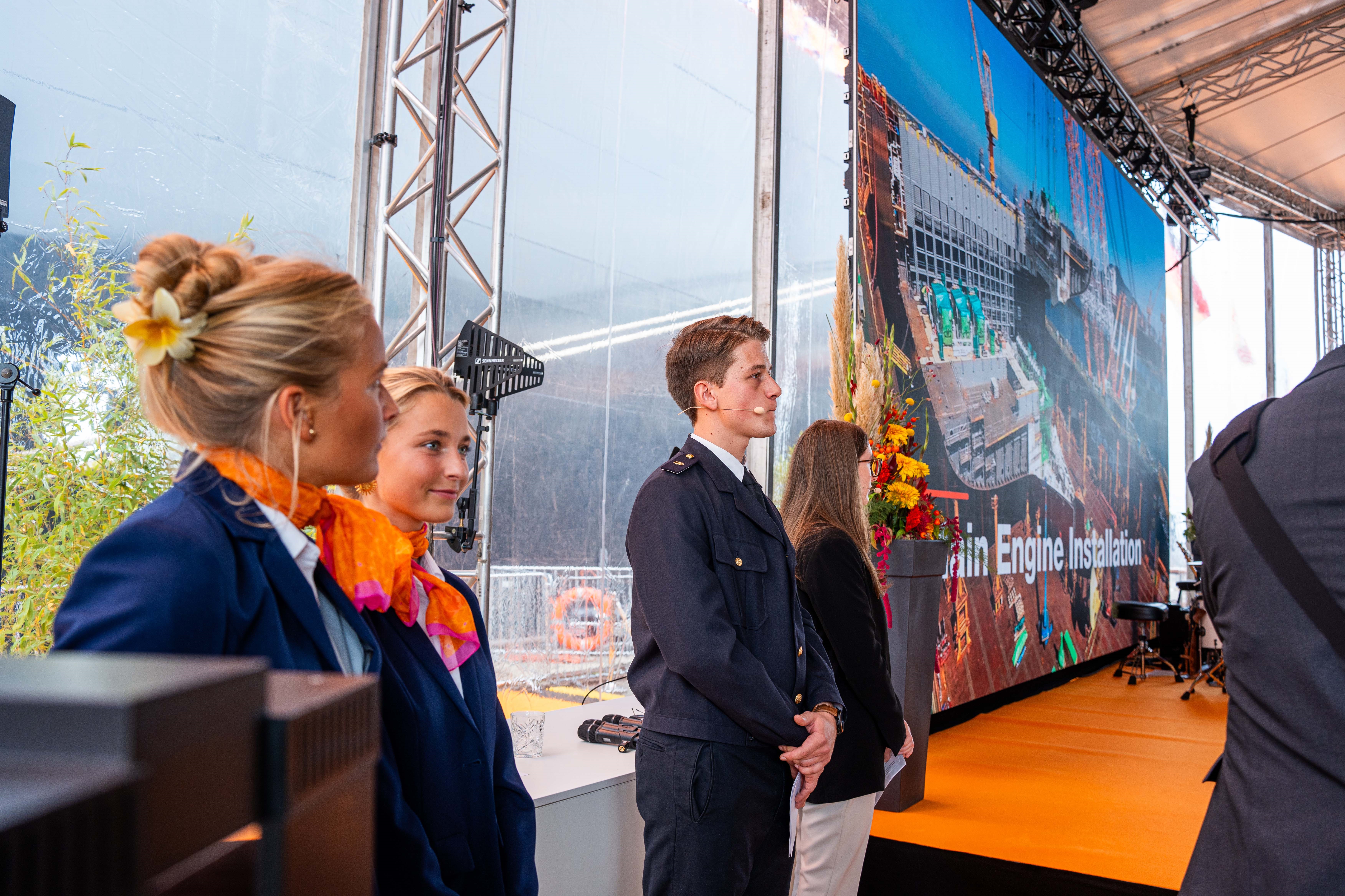 Hosts und Hostessen in eleganten blauen Anzügen mit buntem Halstuch neben einer Bühne, professionelle Organisation und Betreuung bei Messe und Event