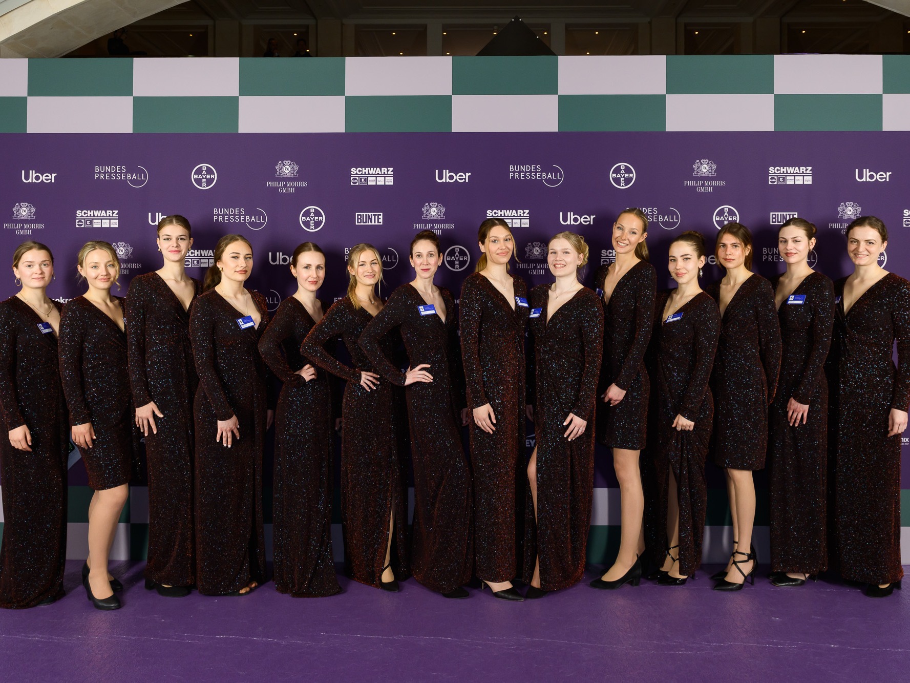 Hostessen in identischen glitzernden Abendkleidern auf lila Teppich bei Gala, eleganter Auftritt mit hohem Beinausschnitt und strahlendem Lächeln