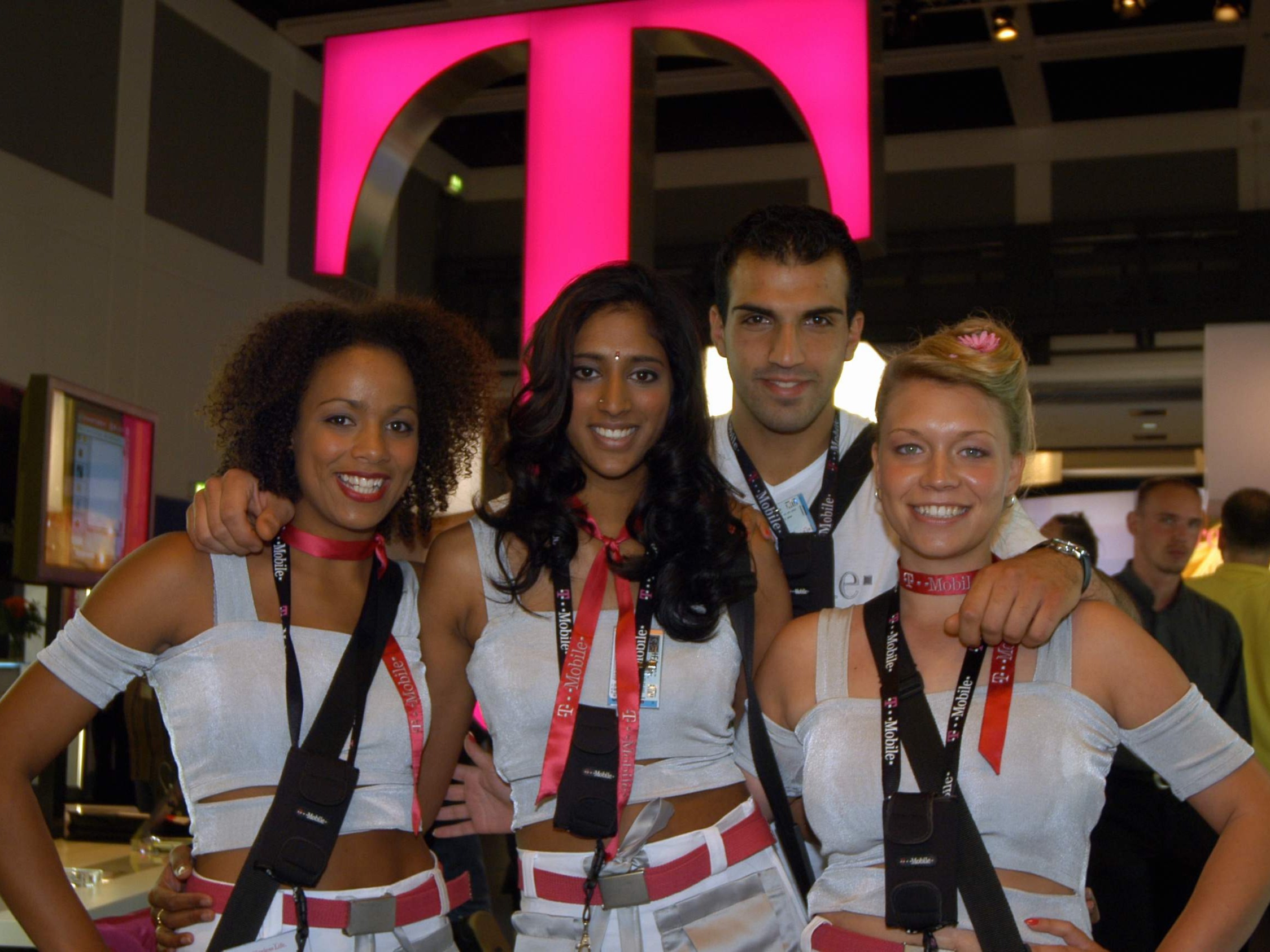 Drei Hostessen und ein Host in weißen Outfits mit auffälligem Gürtel und Badges vor Markenlogo, professioneller Promotion Einsatz bei Messe und Event