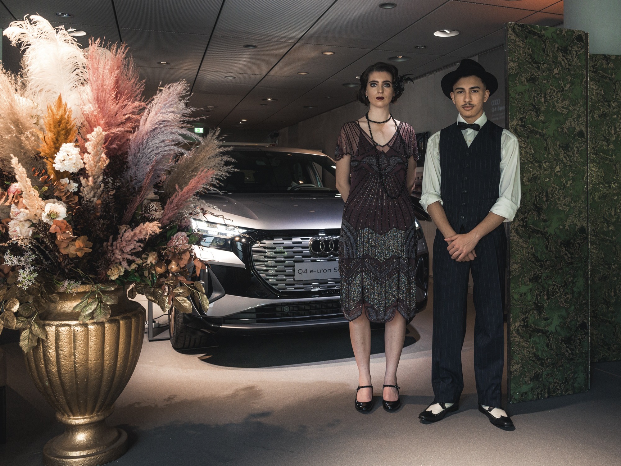 Host und Hostess im 60er-Jahre Look stehen vor einem Auto, ernster Blick in die Kamera, stilvolle Inszenierung bei Event, Shooting und Promotion