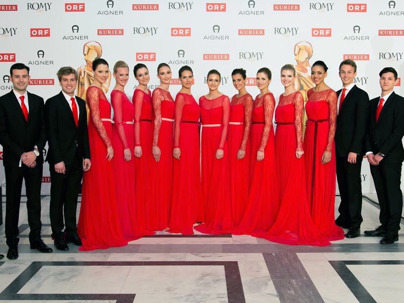 Hostessen in eleganten roten Abendkleidern mit Hochsteckfrisur vor Fotowand, flankiert von Hosts im Anzug, stilvoller Empfang bei Gala und Event