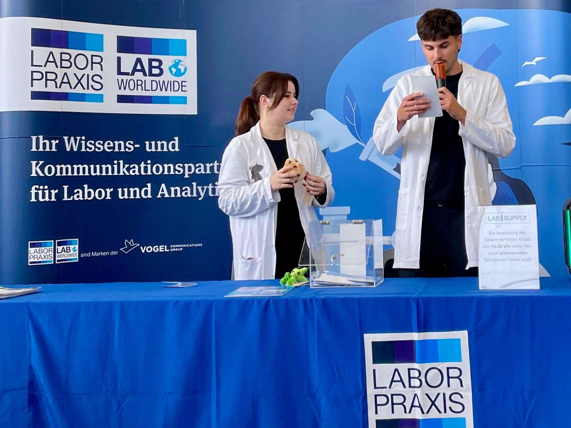 Zwei Personen in wei&szlig;en Kitteln stehen an einem Wissenschaftsstand und lesen Inhalte mit Mikrofon, interaktive Promotion bei Messe und Event