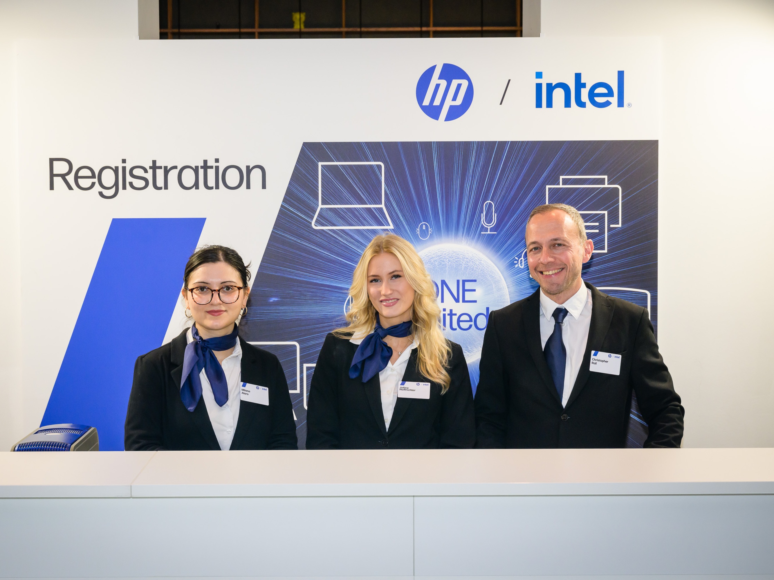Transferhosts und Hostessen in Anzügen mit blauen Halstüchern und Krawatte empfangen Gäste am Tresen, professioneller Shuttle- und Empfangsservice bei Event
