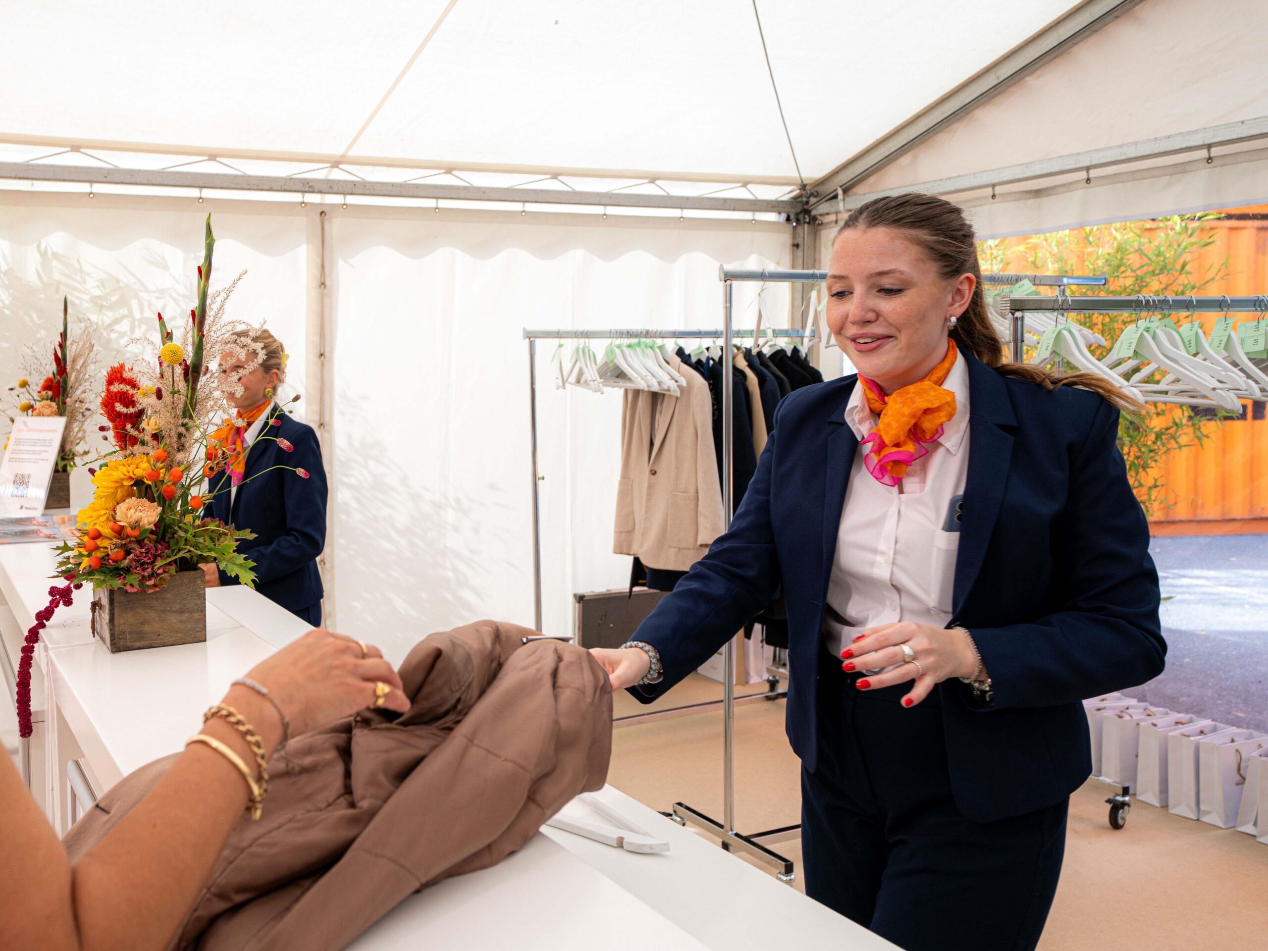Hostess im eleganten Hosenanzug mit orangem Halstuch nimmt an der Garderobe eine Jacke entgegen, professioneller Empfangsservice bei Event und Gala.