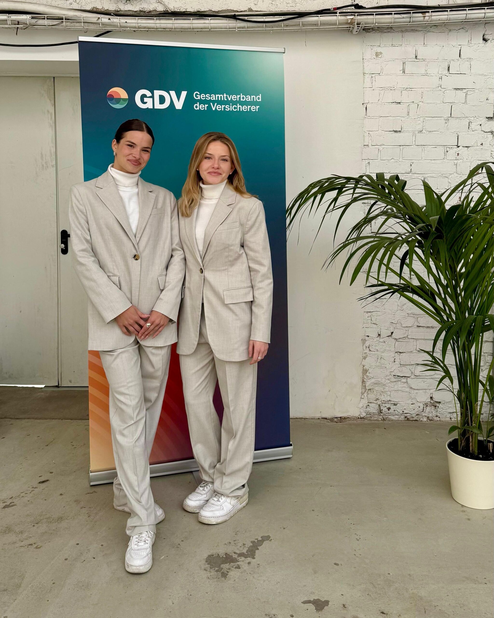 Zwei Hostessen in eleganten grauen Hosenanzügen stehen vor einem Messeposter, professioneller Auftritt bei Event, Konferenz und Markenpräsentation