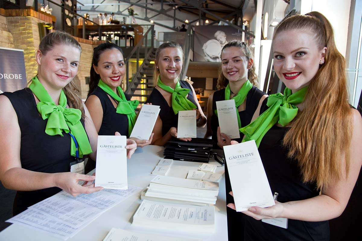 > Hostessen in schwarzen Kleidern mit grünem Halstuch an der Akkreditierung zeigen ein Dokument in die Kamera, professioneller Check-in bei Event und Messe