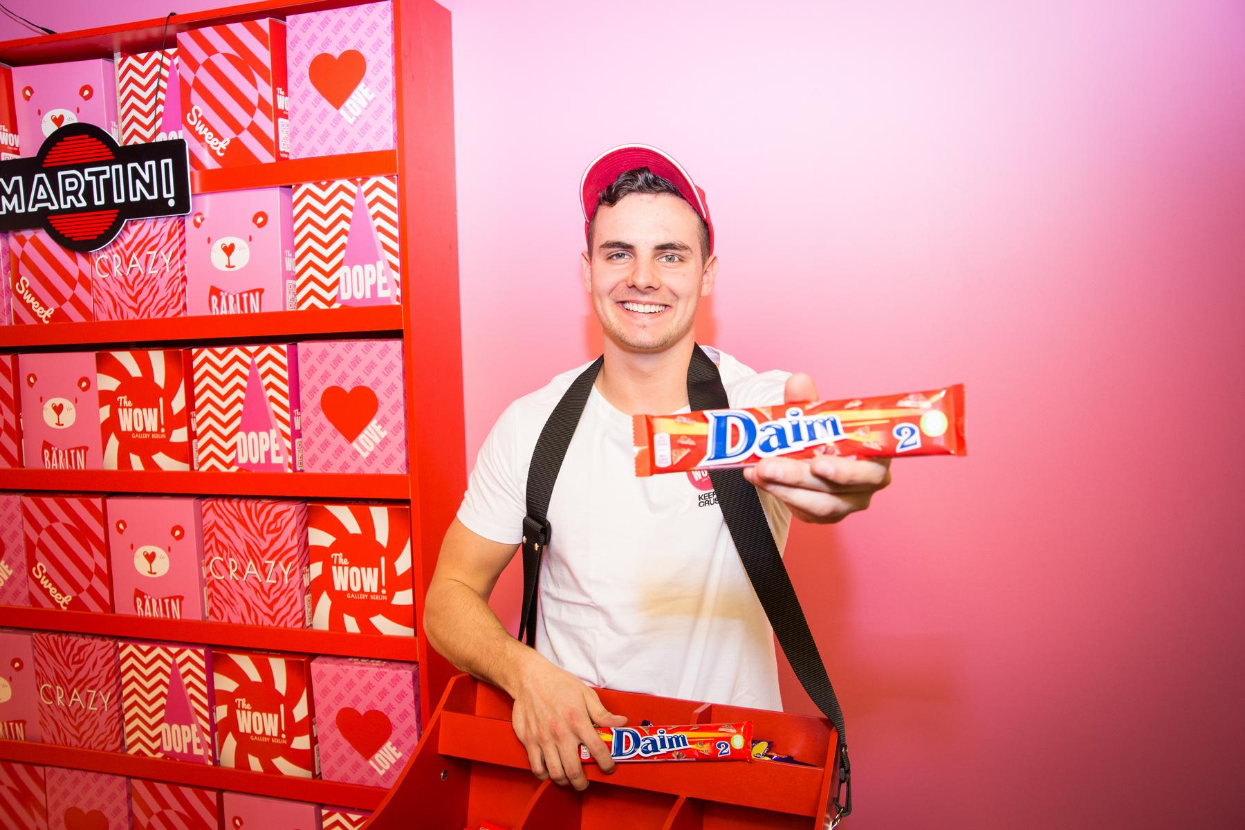 Promoter zeigt einen Daim Riegel in die Kamera, trägt eine Verteilbox um den Hals vor roter Markenwelt, aufmerksamkeitsstarke Promotion Aktion bei Event