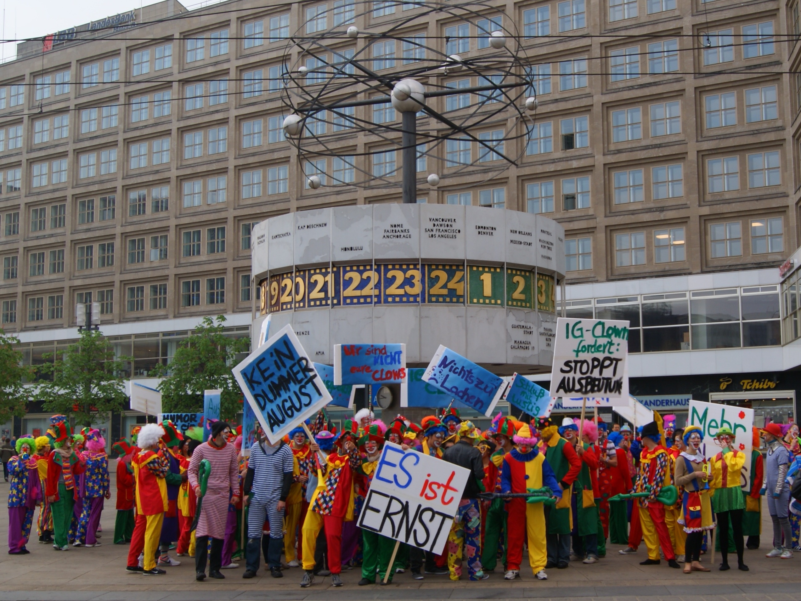Viele Personen in Clown-Kostümen mit Schildern vor der Weltuhr am Alexanderplatz, aufmerksamkeitsstarke Promotion Aktion in Berlin bei Event und Kampagne