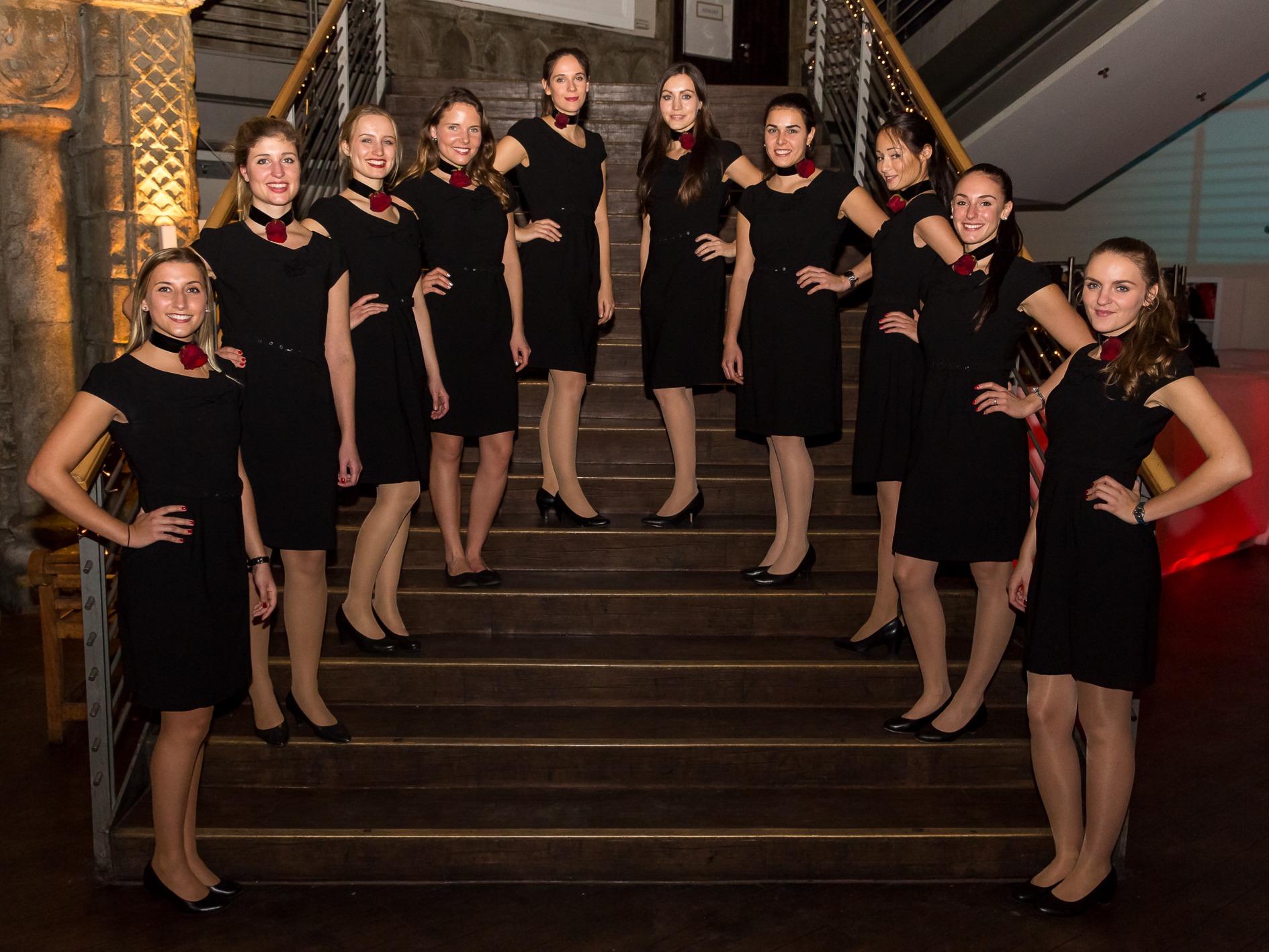 Zehn Eventhostessen in schwarzen Business-Outfits mit Choker und roter Rose stehen auf einer Treppe, eleganter Auftritt bei Event und Promotion Einsatz