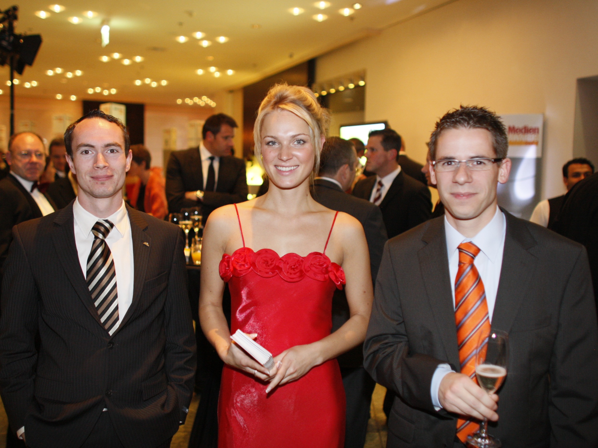 > Eventhostess im roten Abendkleid mit Dutt steht zwischen zwei Gästen im Anzug, eleganter Empfang und stilvolle Betreuung bei Gala und Event