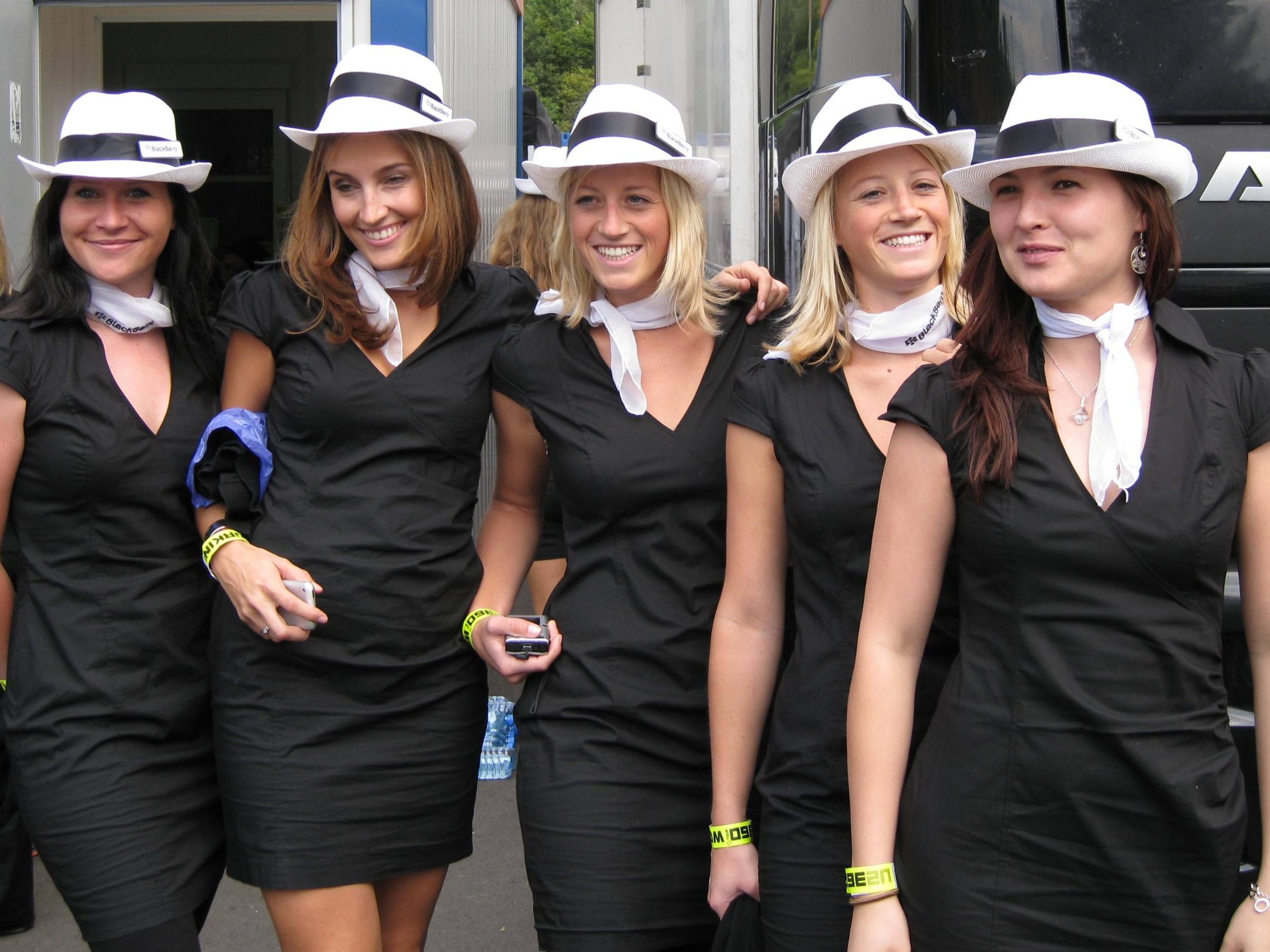 Fünf Hostessen in schwarzen Kleidern mit weißen Hüten und Halstüchern auf einer Messe, unterwegs im Einsatz bei Promotion und Eventbetreuung