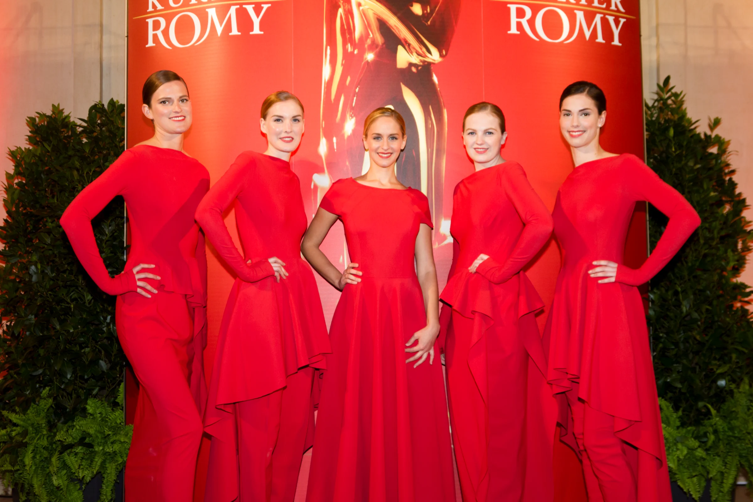 Fünf Modelhostessen in roten Abendkleidern posieren auf dem roten Teppich, eleganter Auftritt mit Lächeln bei Gala, Event und Markenpräsentation