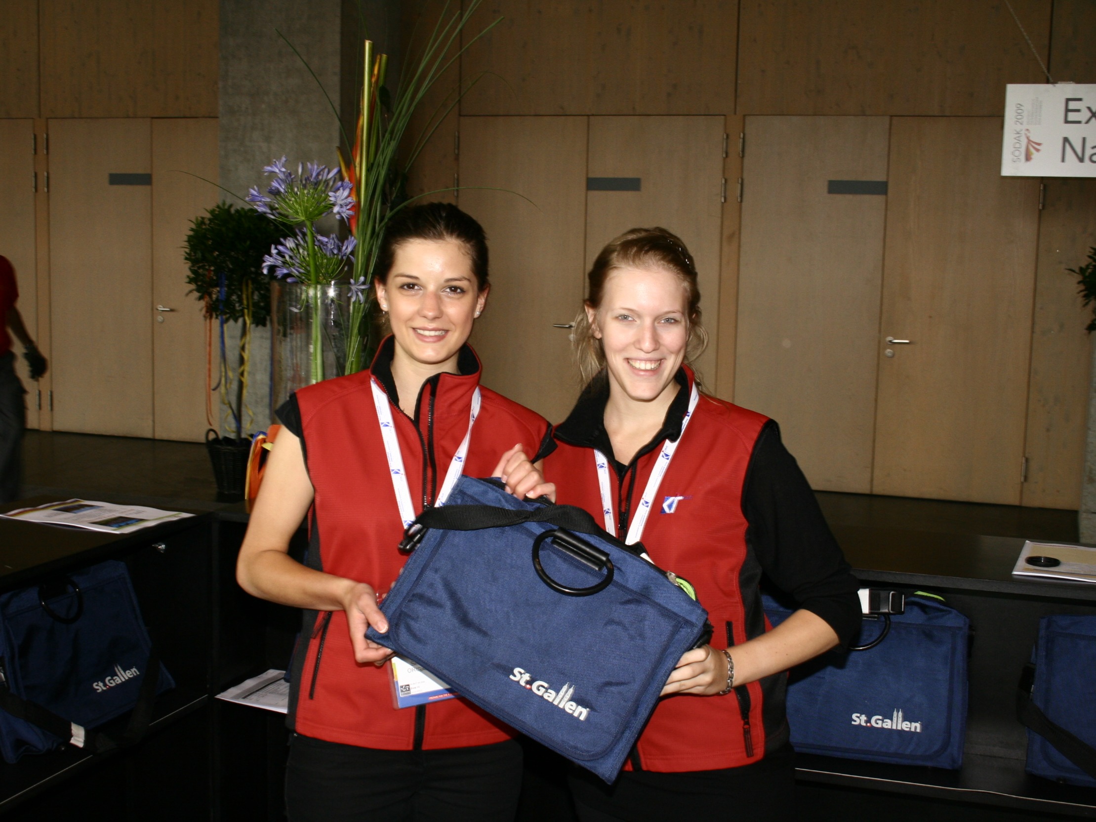 Zwei Hostessen in identischen Outfits halten gemeinsam eine blaue Tasche und lächeln in die Kamera, Promotion Einsatz auf einer Messe mit Markenpräsentation