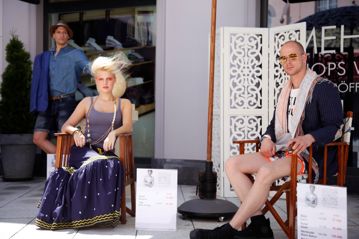 > Modelhost und Modelhostess sitzen vor einem Schaufenster im Fotoshooting-Setting, auffällige Outfits und direkter Blick in die Kamera bei Promotion und Fashion Event