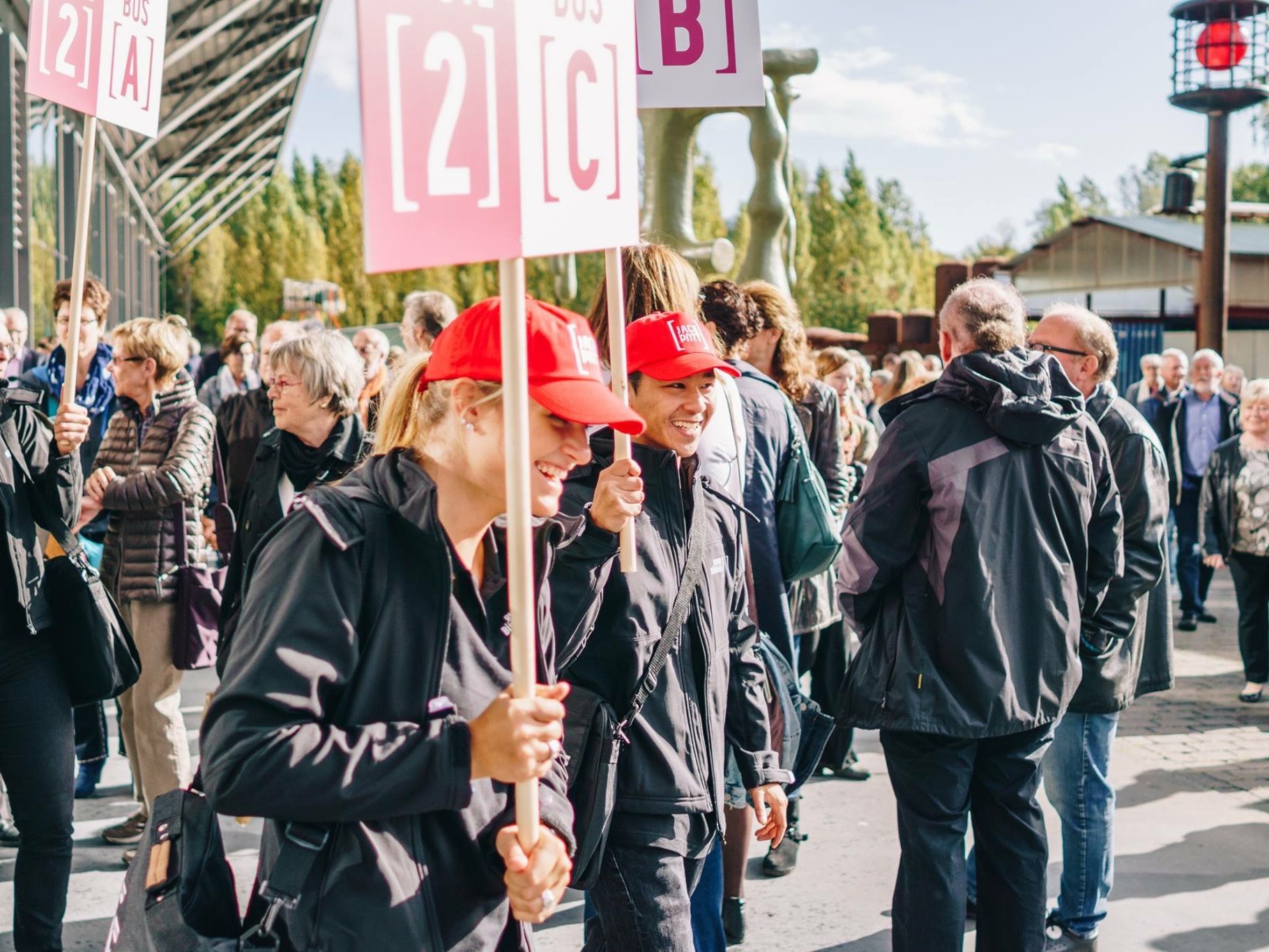 Hostess mit „2C“ Schild, roter Cap und Regenjacke begrüßt Besucher, im Hintergrund weiteres Promotion Team im Einsatz bei Event und Besucherlenkung