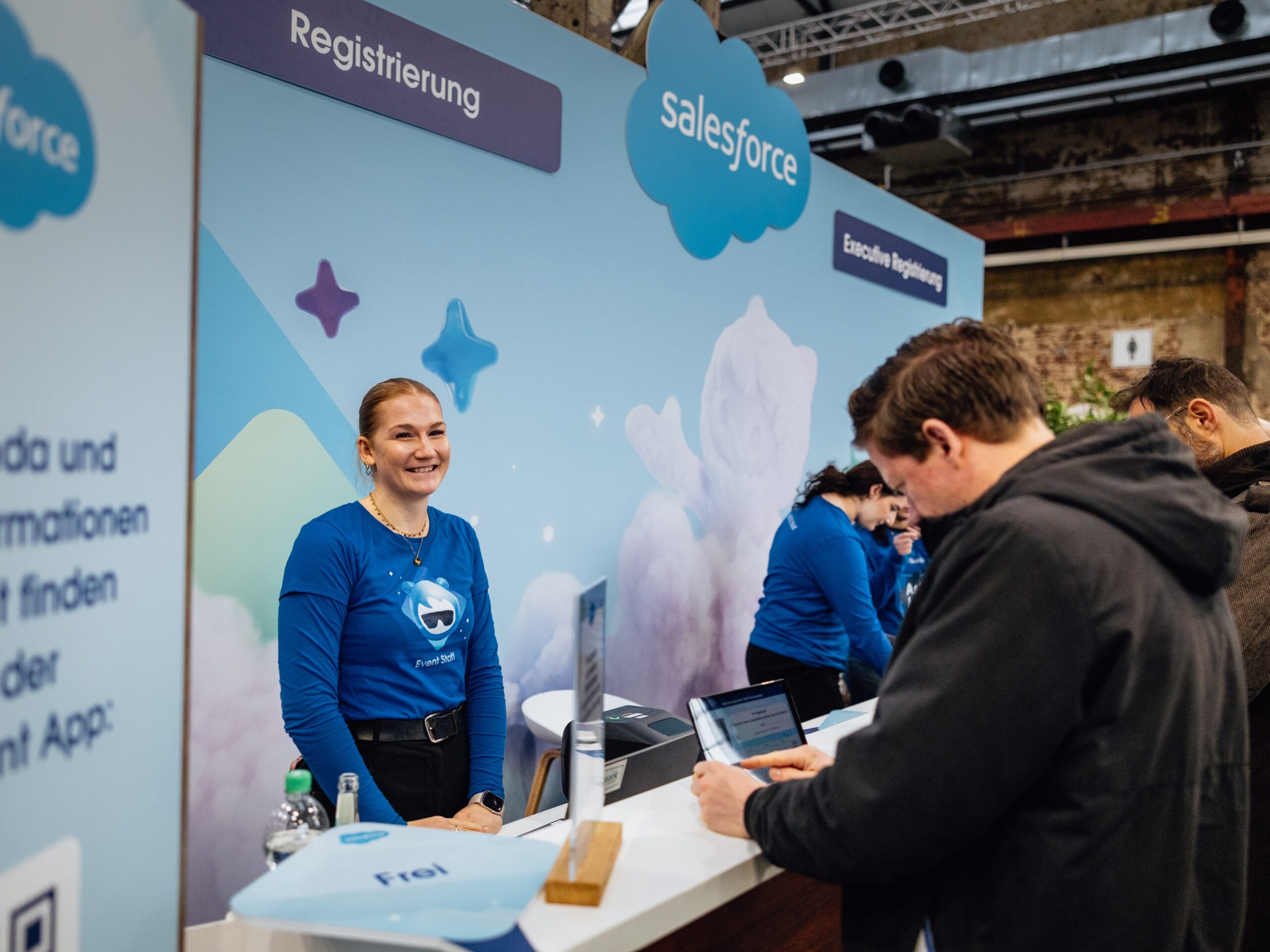 > Messehostess mit blauem Pullover und Dutt betreut einen Gast hinter einem Messestand, professioneller Service bei Messe, Event und Markenpräsentation