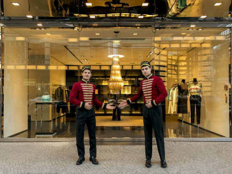 Zwei Hosts und Hostessen in Pagen-Outfits vor einer gl&auml;sernen T&uuml;r, im Hintergrund ein leuchtender Kronleuchter, eleganter Empfang bei Event und Gala