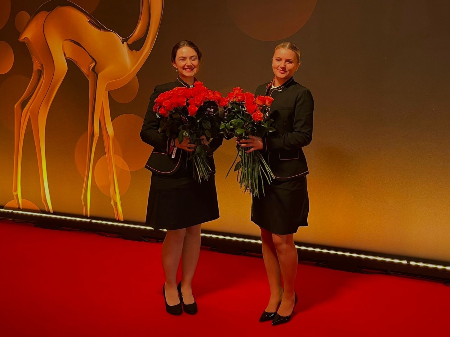 Hostessen von cinnamon in schwarzen Business-Outfits mit knielangen Röcken und roten Rosen auf dem roten Teppich, eleganter Empfangsservice bei Preisverleihung und Gala Event