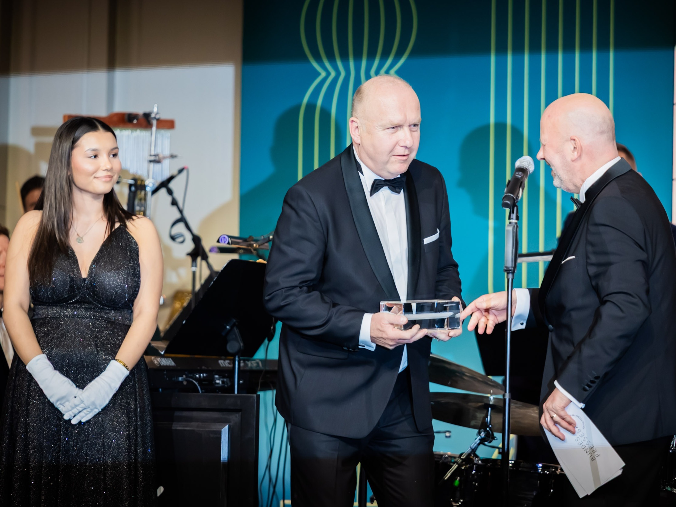 Bühnenhostess beim Bundespresseball im glitzernden Abendkleid mit weißen Handschuhen neben Gästen im Smoking, eleganter Gala-Moment bei Preisverleihung und Event