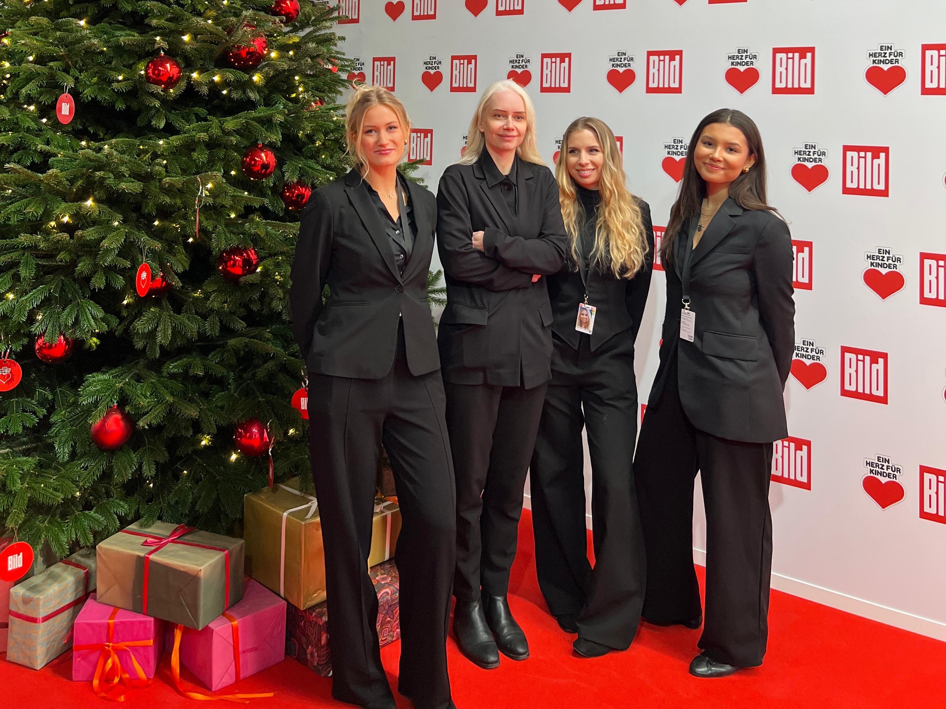 Hostessen in schwarzen Outfits vor einer Fotowand neben einem Weihnachtsbaum, eleganter Empfangsservice bei Weihnachtsfeier, Gala und Event