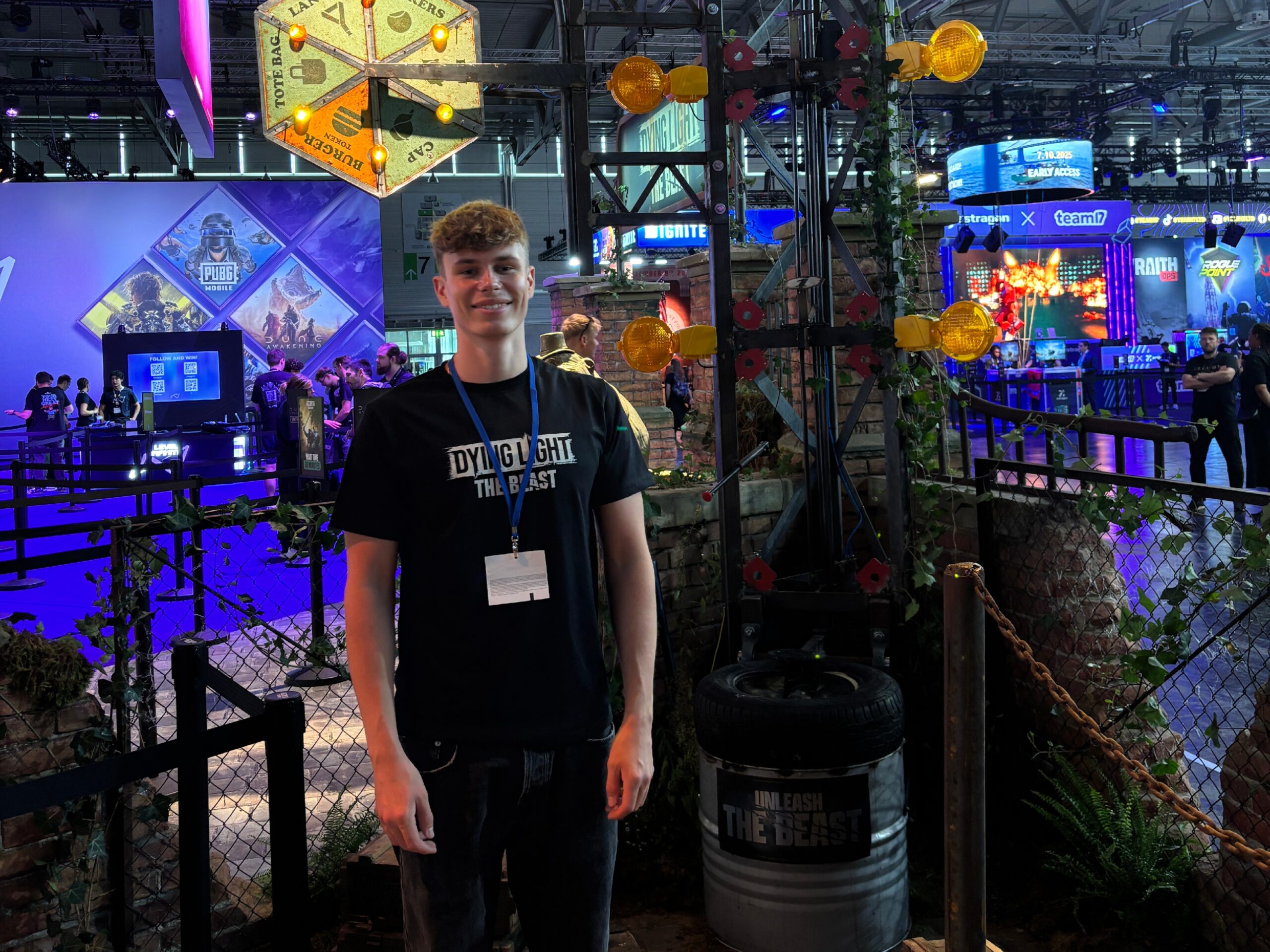 Host auf der gamescom vor einer Attraktion in schwarzem Outfit mit bedrucktem T-Shirt, lächelnder Promotion Einsatz bei Gaming-Event und Markenpräsentation