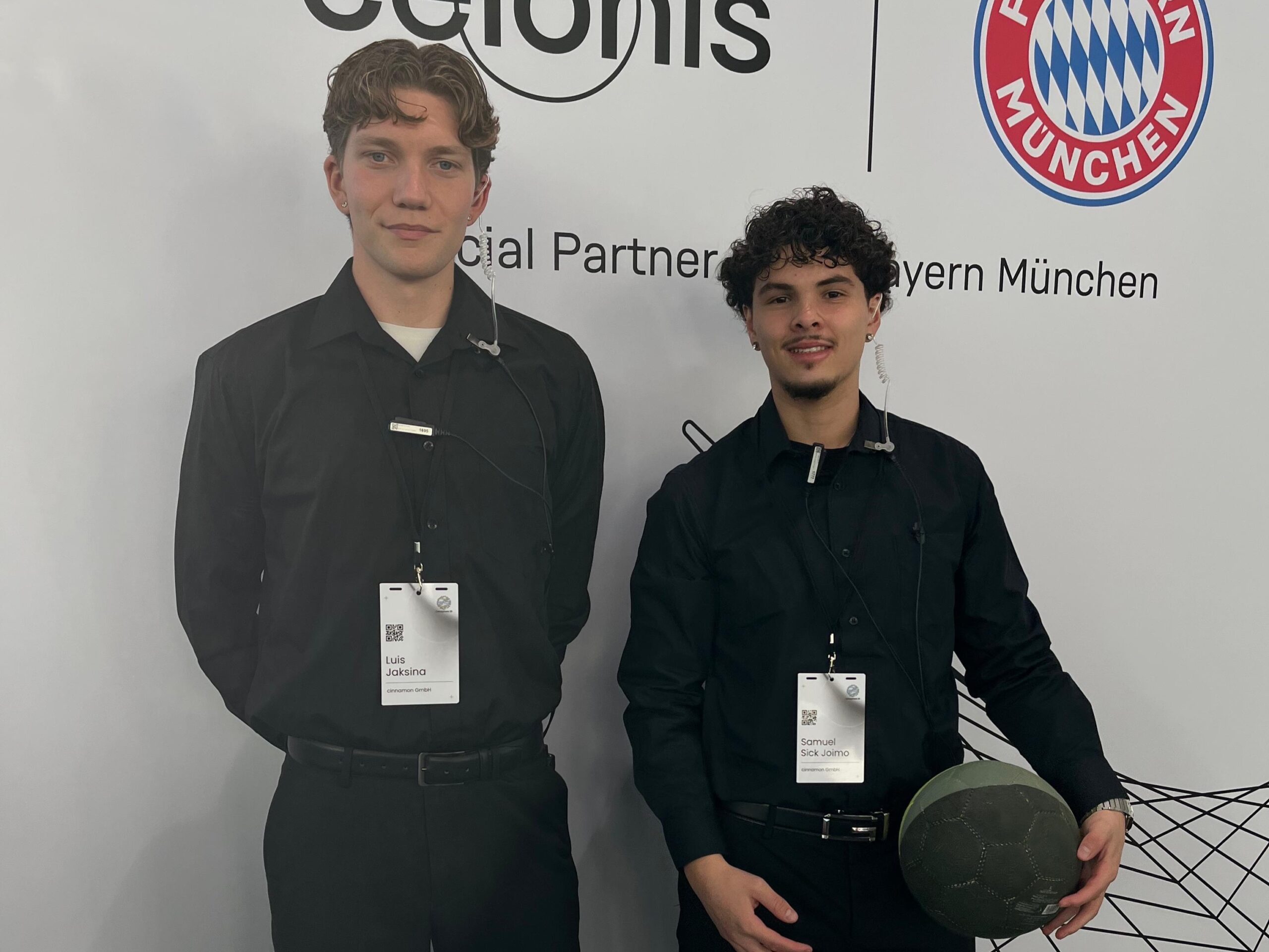 Zwei Hosts bei einem Bayern München Event vor einer Fotowand, einer hält einen Fußball in der Hand, sportliche Promotion und Markenpräsentation bei Veranstaltung
