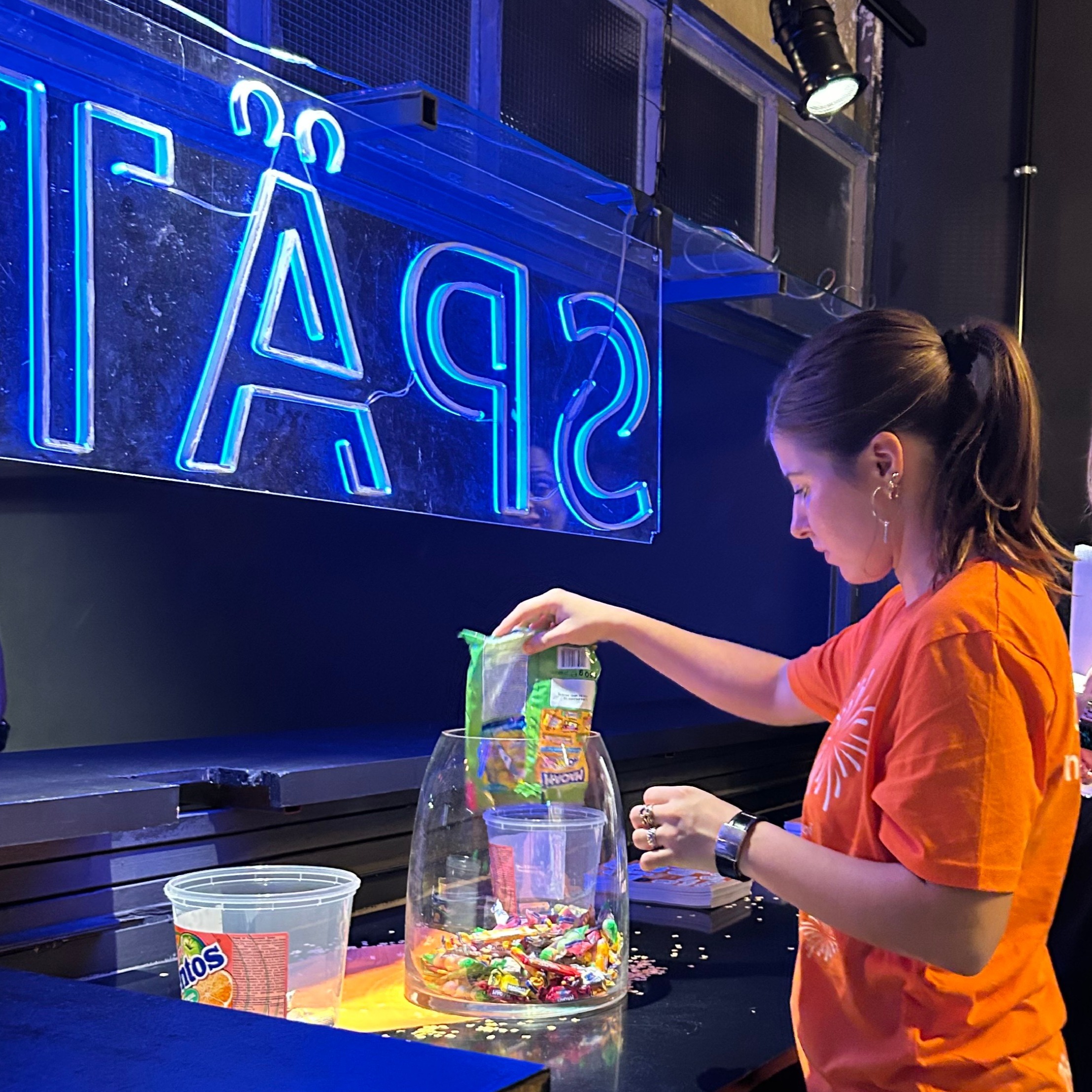 Hostess im orangenen T-Shirt füllt Süßigkeiten in ein Glas, freundlicher Promotion Einsatz bei Event, Messe und Markenaktion
