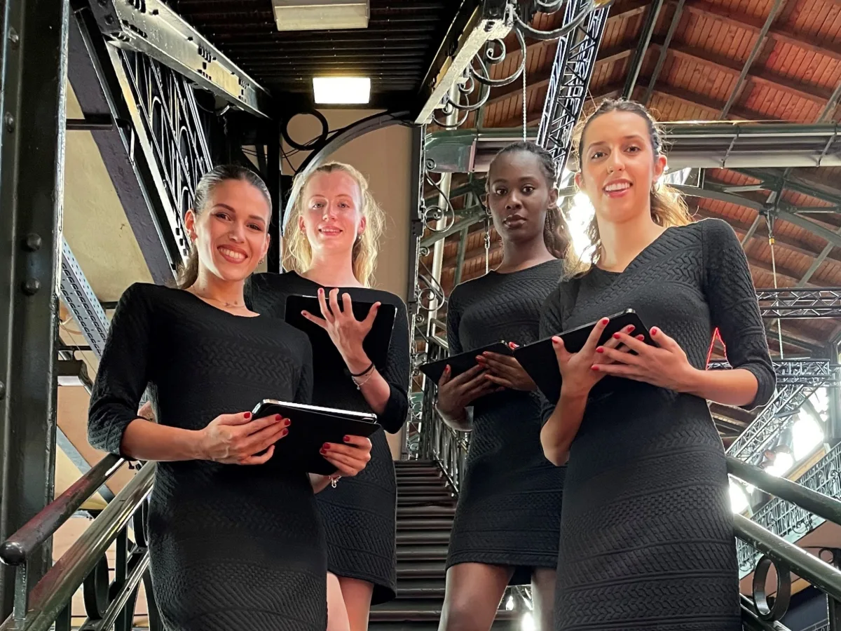 Vier Hostessen in kleinen schwarzen Kleidern mit Tablets in der Hand stehen auf einer Treppe, professioneller Empfangsservice bei Event, Messe und Gala