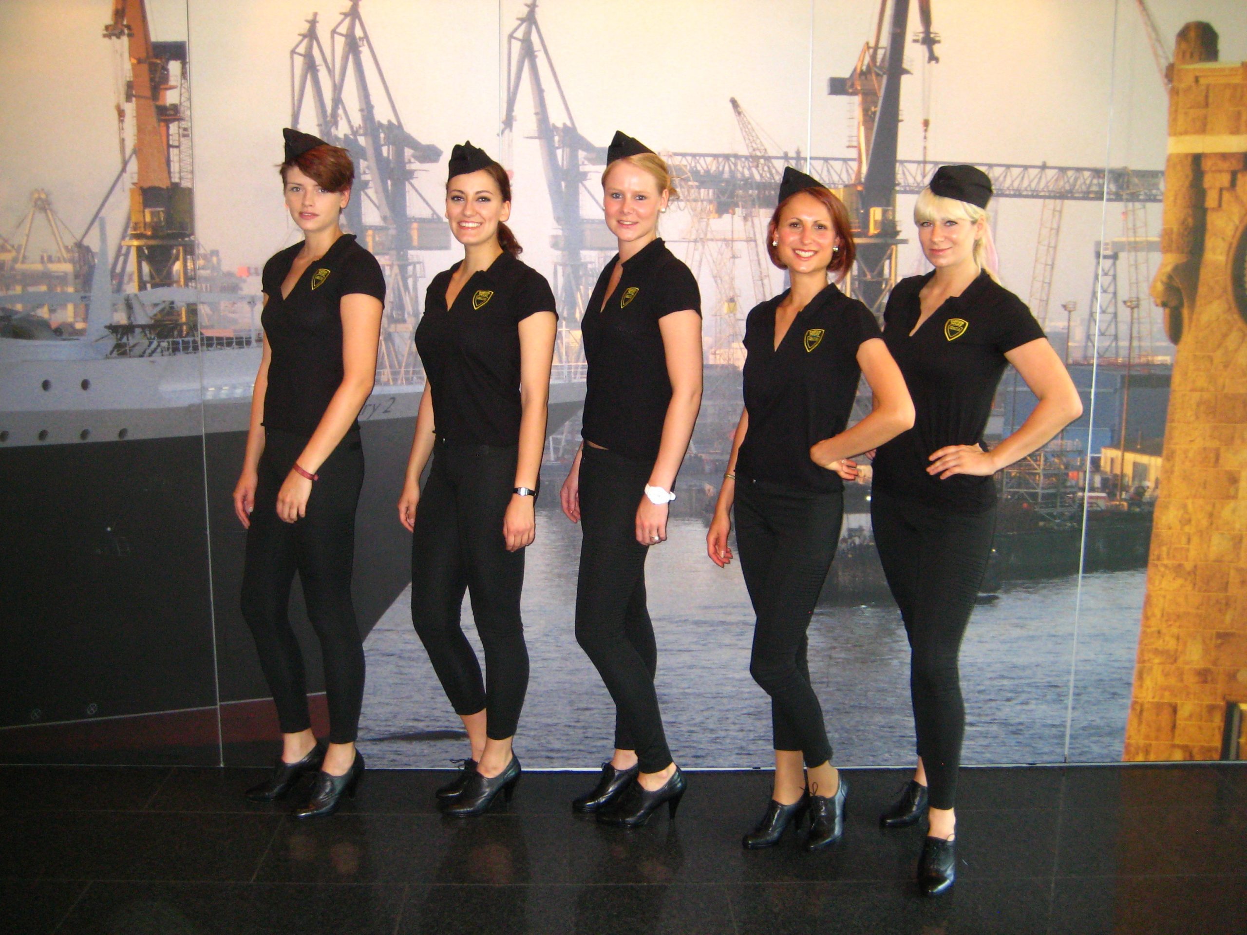Hostessen in All-Black Outfits mit schwarzen Stewardessenkappen vor einer Fotowand mit Hafenmotiv, eleganter Promotion Einsatz von cinnamon bei Event und Markenpräsentation