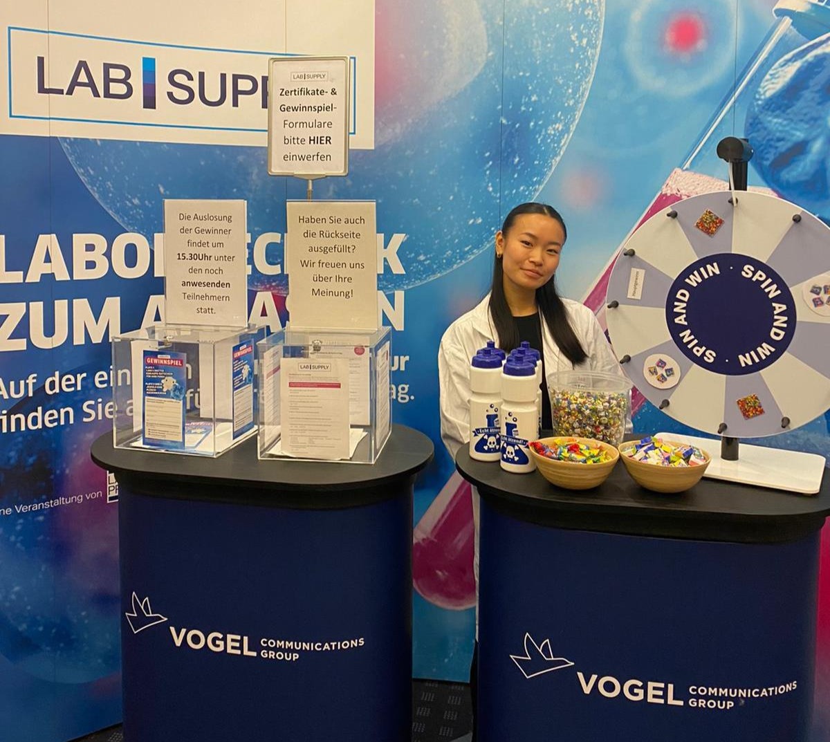 Messehostess im Kittel auf der Lab Supply Messe in Frankfurt mit Flyern und Glücksrad, lächelnde Markenrepräsentation bei Event und Promotion Aktion