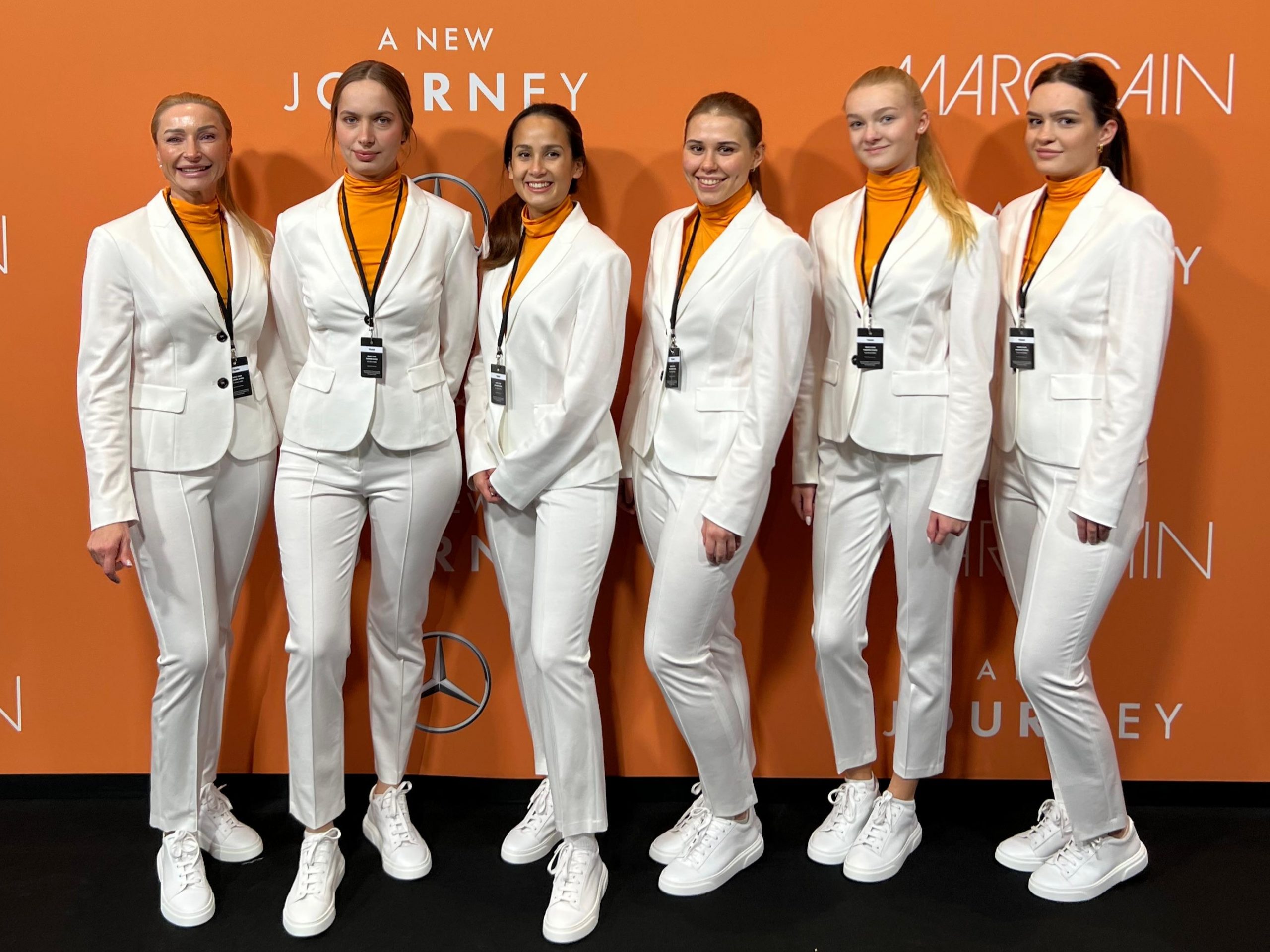 Hostessen in weißen Anzügen mit orangenen T-Shirts vor einer orangenen Fotowand, lächelnder Promotion Einsatz mit Badges und weißen Sneakern bei Event und Markenpräsentation