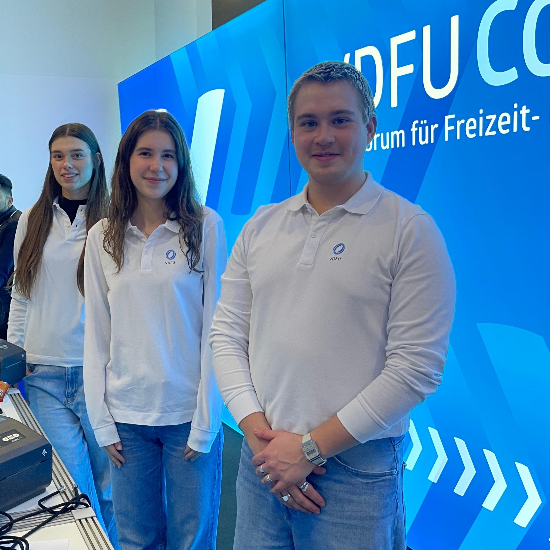 Hosts und Hostessen in weißen Longsleeves und blauen Hosen vor einer Fotowand auf einer Messe in Frankfurt, lächelnder Akkreditierungs- und Empfangsservice bei Event und Konferenz.
