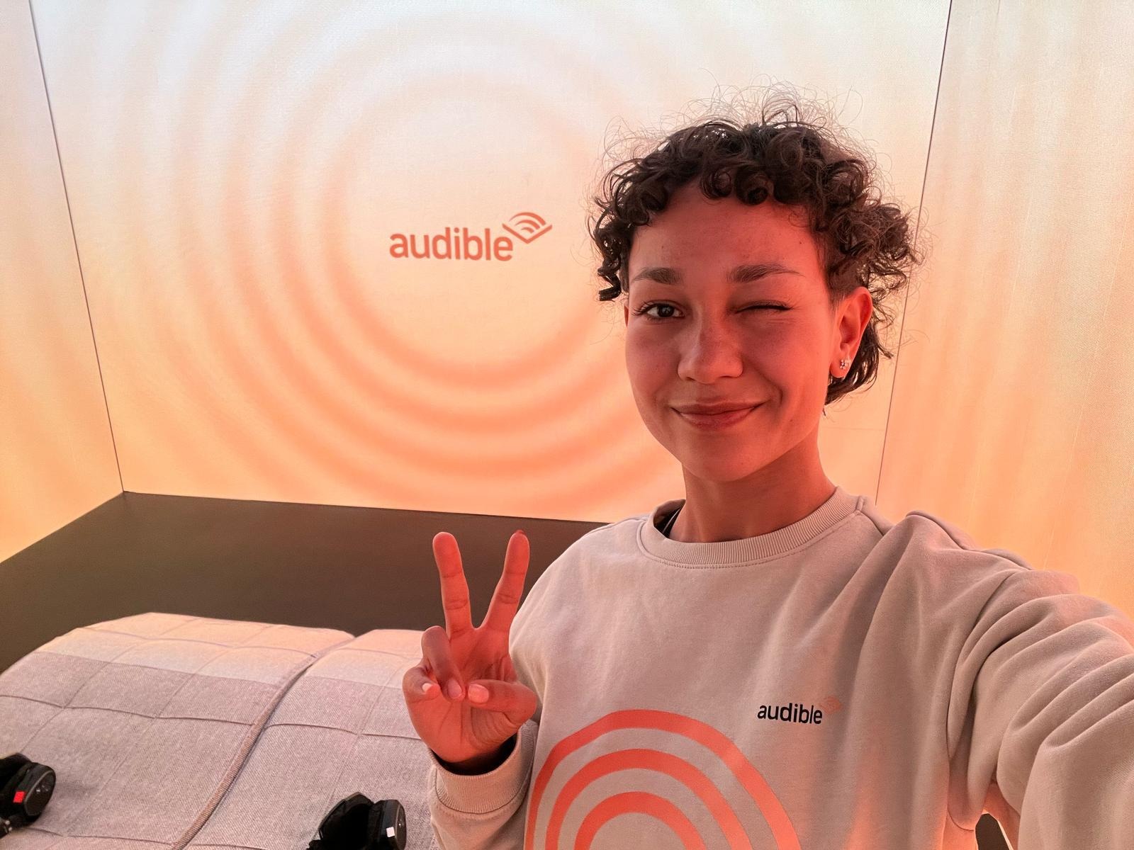 Promoterin in Dortmund vor einem Audible Schild mit grauem Audible Hoodie zeigt ein Peace-Zeichen, freundliche Markenpromotion bei Event und Sampling Aktion
