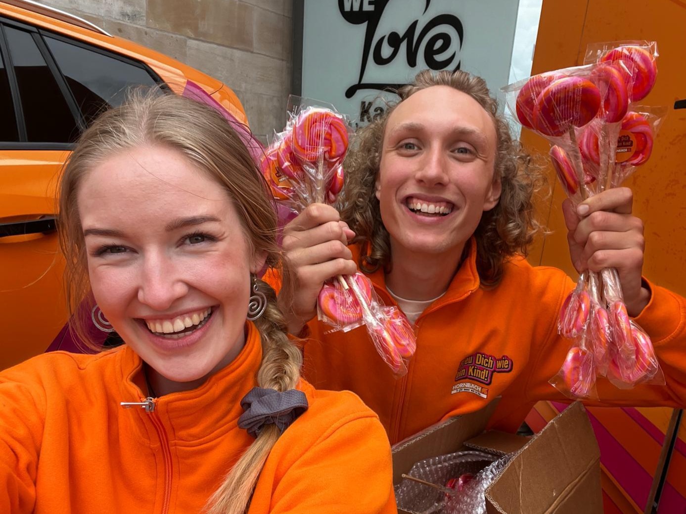 Promoter in orangener Hornbach Kleidung halten Goodies in die Kamera und lächeln, auffällige Promotion Aktion und Markenaktivierung bei Event und Sampling Einsatz