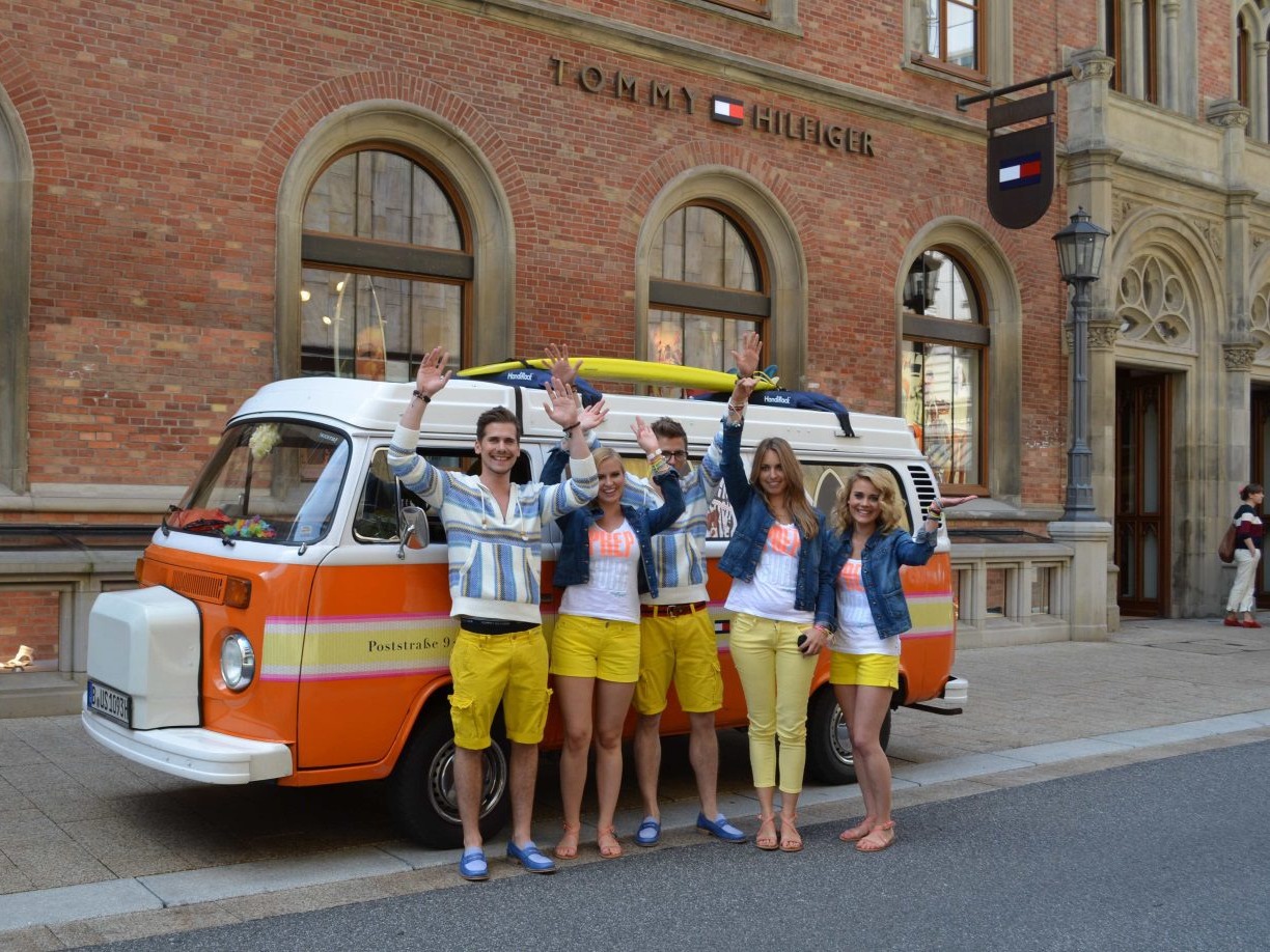 Promoter und Promoterinnen in schrillen Outfits vor einem Van jubeln mit erhobenen Händen, fröhliche Promotion Aktion bei Event und Markenaktivierung