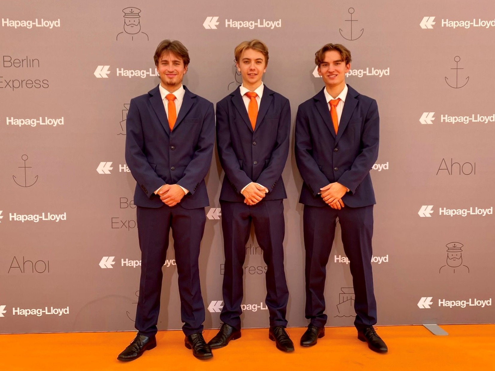 Drei Hosts in blauen Anzügen mit orangenen Krawatten stehen vor einer Fotowand, eleganter Empfangsservice und professioneller Eventauftritt bei Promotion und Gala