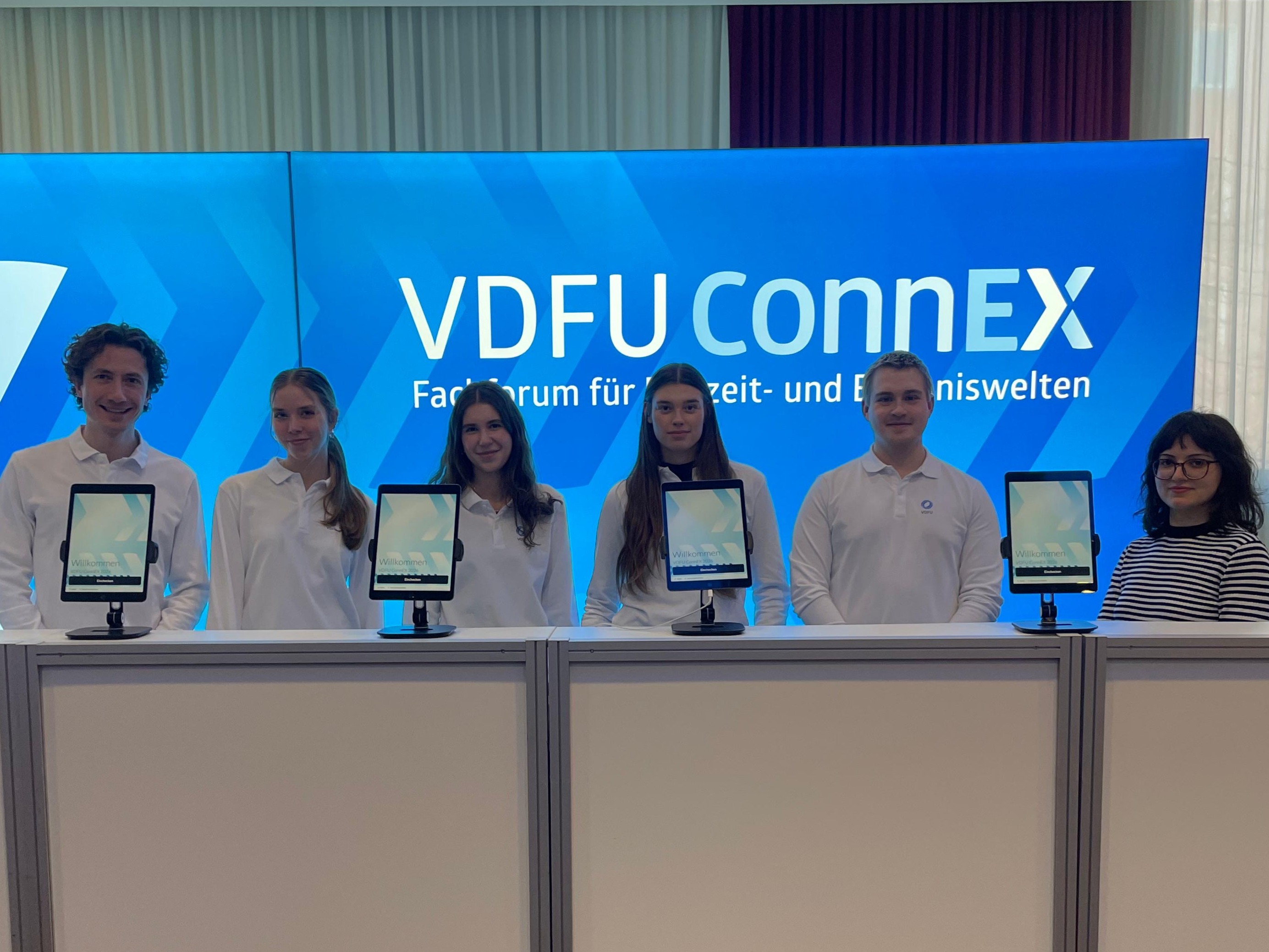 Sechs Hosts und Hostessen in weißen Longsleeves an der Akkreditierung vor blauer Fotowand in Freiburg, professioneller Check-in und Gästeservice bei Event und Messe
