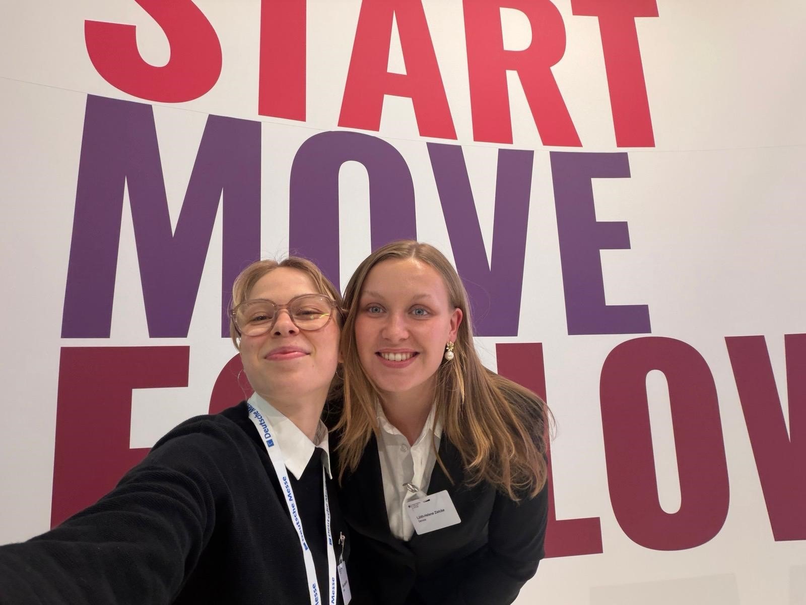 Zwei Messehostessen machen ein Selfie vor einer Fotowand, lächelnder Promotion Einsatz in weißen Hemden und schwarzen Sweatshirts bei Event und Messe