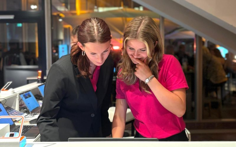 Zwei Hostessen in pinken Oberteilen, eine mit Blazer, stehen hinter einem Tresen und schauen gemeinsam auf einen Bildschirm, konzentrierte Betreuung bei Event und Messe