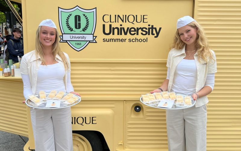 Zwei Promotion Hostessen stehen vor einem gelben Retro-Bus mit Markenbranding, präsentieren Produkte auf Tablets, lächelnder Einsatz bei Event und Markenaktion