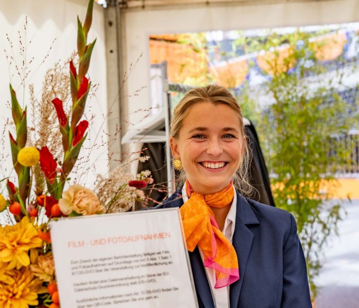 Hostess im blauen Hosenanzug mit orangem Halstuch arbeitet an der Garderobe, lächelnder Empfangsservice bei Event, Gala und Veranstaltung