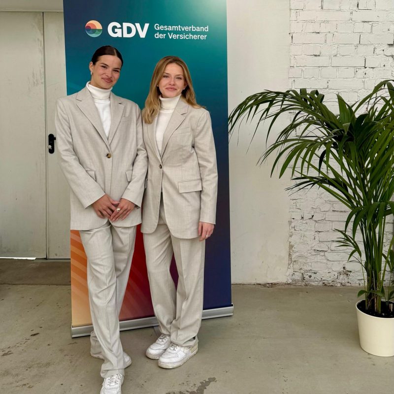 Zwei Hostessen in eleganten grauen Hosenanzügen stehen vor einem Messeposter, professioneller Auftritt bei Event, Konferenz und Markenpräsentation