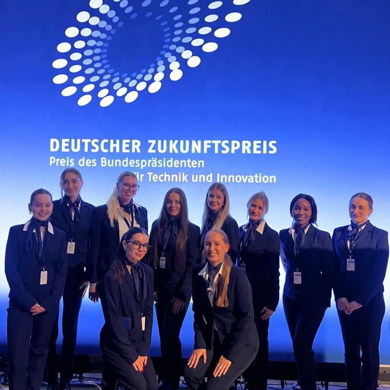 Hostessen in blauen Hosenanzügen mit passendem Halstuch vor einer Fotowand beim Deutschen Zukunftspreis, professioneller Empfangsservice bei Gala und Event